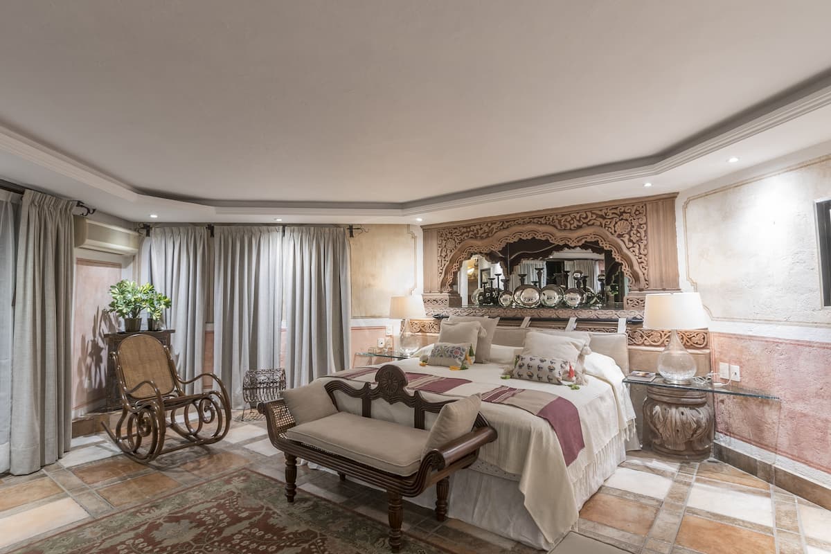 A spacious bedroom with a large bed, a sitting area with a chair and a small table, and a decorative mirror above the bed.