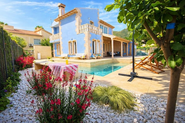 A two-story house with a pool, surrounded by a garden with pebbles and flowers.