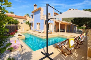 Une piscine entourée de chaises longues et de parasols, elle-même bordée d’une maison avec un toit en tuiles et une pergola.