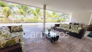 A spacious outdoor seating area with a glass table, wicker furniture, and a view of a landscaped garden with palm trees. - Palm Village Acqua (Ipojuca)
