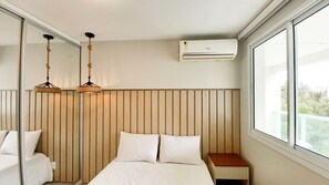 A hotel room with a bed, bedside tables, a hanging light fixture, a window, and an air conditioning unit.
