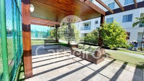A covered outdoor area with a bench, a wicker sofa, and a tree. - Palm Village Acqua (Ipojuca)