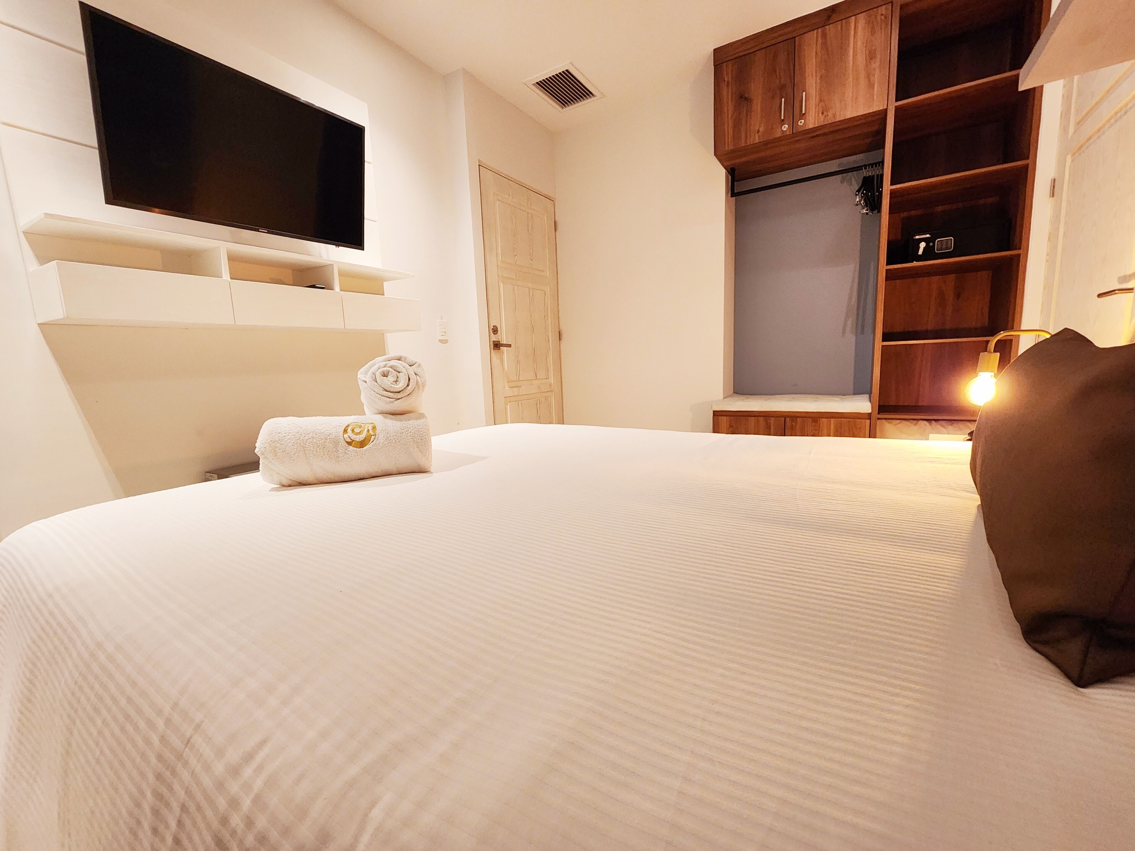 A hotel room with a large bed, a flat-screen TV mounted on the wall, wooden shelving, and a wardrobe.
