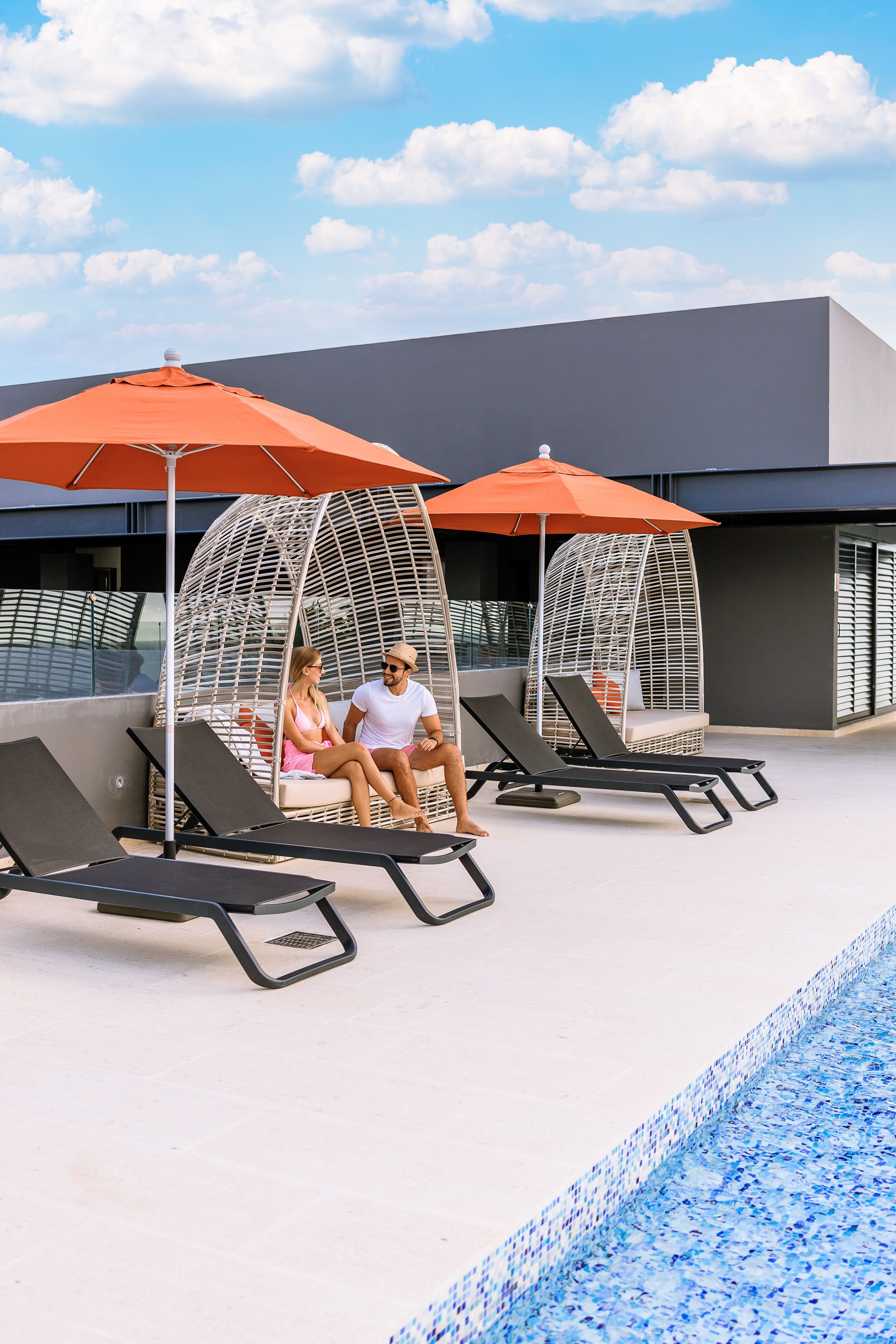 A poolside area with lounge chairs and umbrellas, a modern building in the background, and a clear sky.
