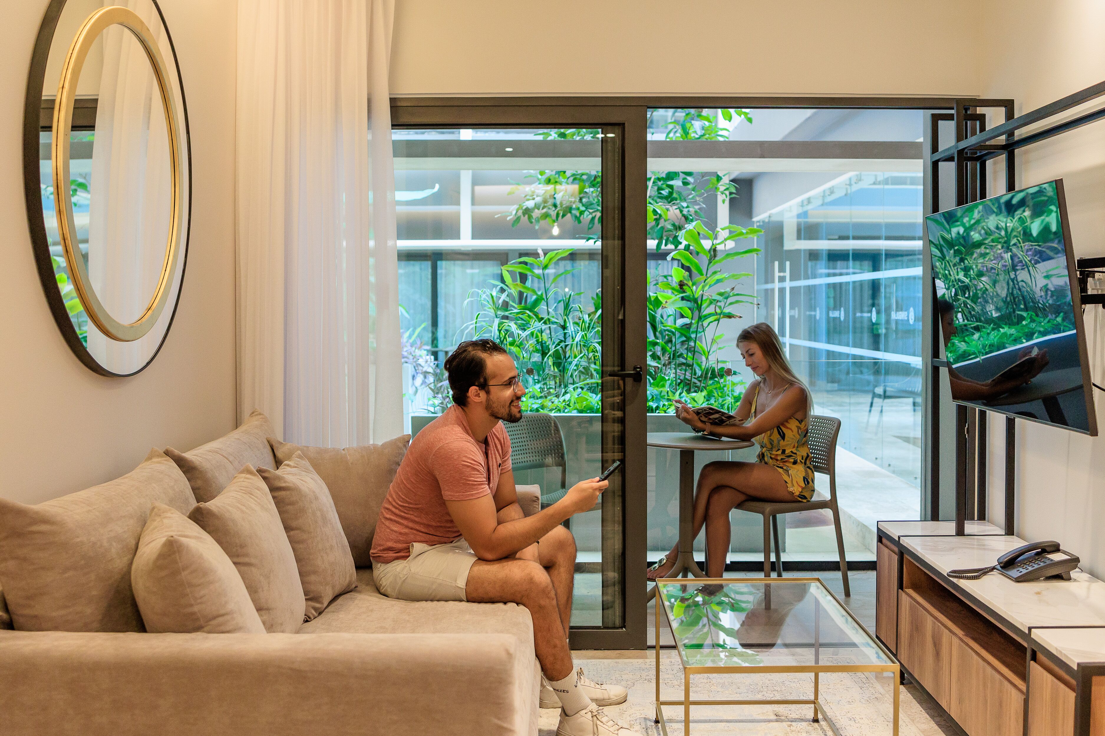 A modern living room with a sofa, a glass coffee table, a TV, and a glass door leading to a balcony with plants.