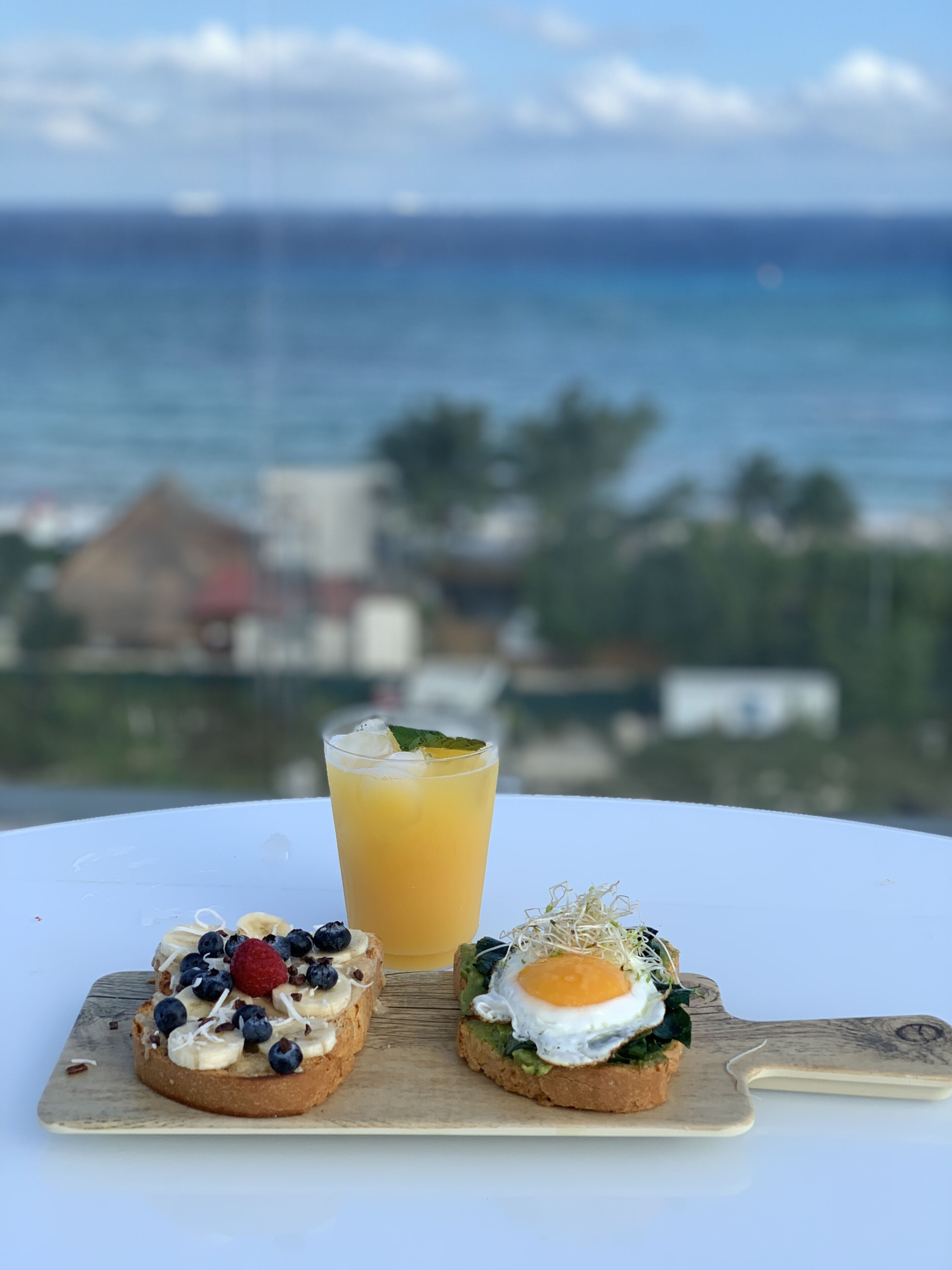 Et bord med to morgenmadsretter og et glas appelsinjuice, med udsigt til havet og bygninger.