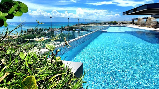 A clear blue pool with a glass barrier, surrounded by tropical plants and overlooking a coastal view. - Singular Dream Beach Residences (Playa del Carmen)