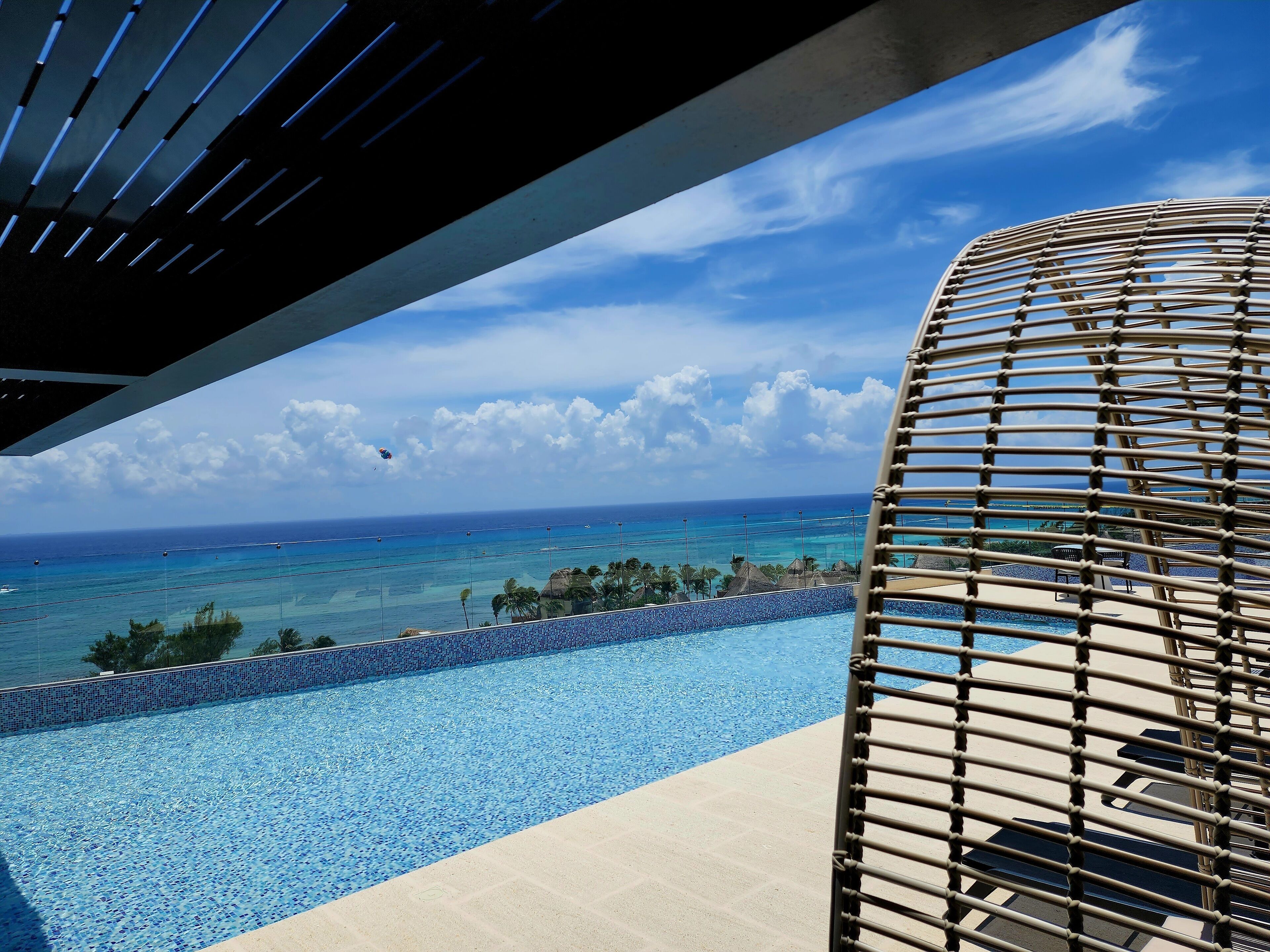 Blick auf einen Pool, einen klaren blauen Himmel, das Meer und einen Strand mit Palmen.