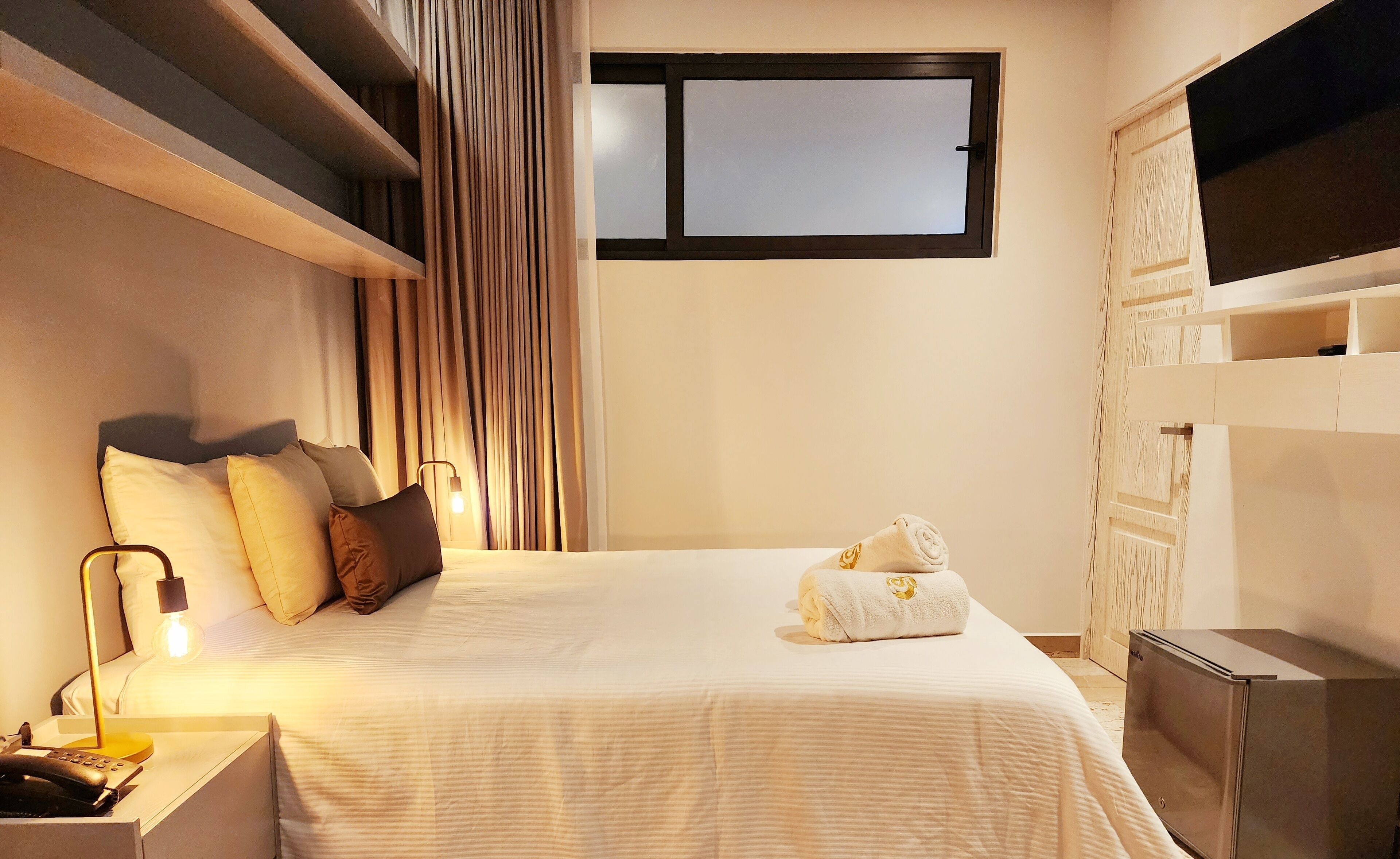A hotel room with a bed, a TV mounted on the wall, a desk with a lamp, and a window with curtains.