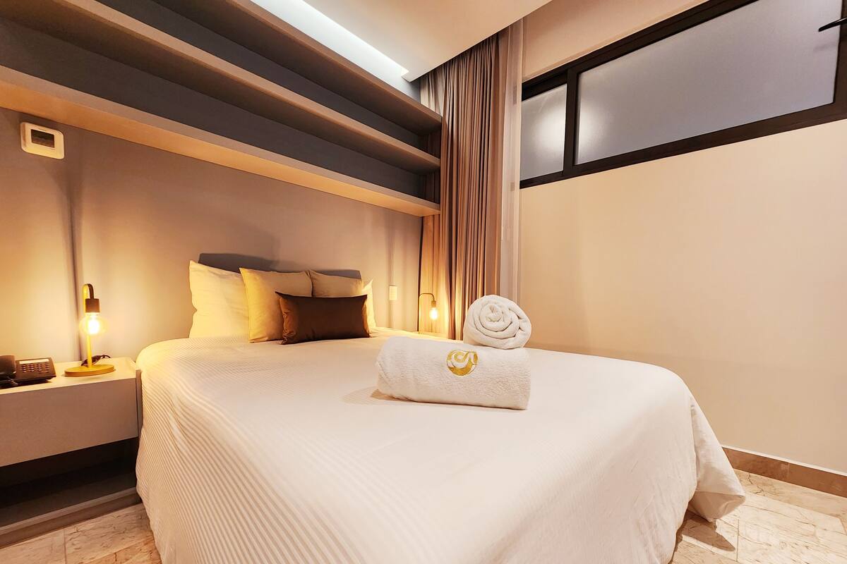 Hotelkamer met een groot bed, nachtkastje met een lamp, telefoon en een opgerolde handdoek met een logo.