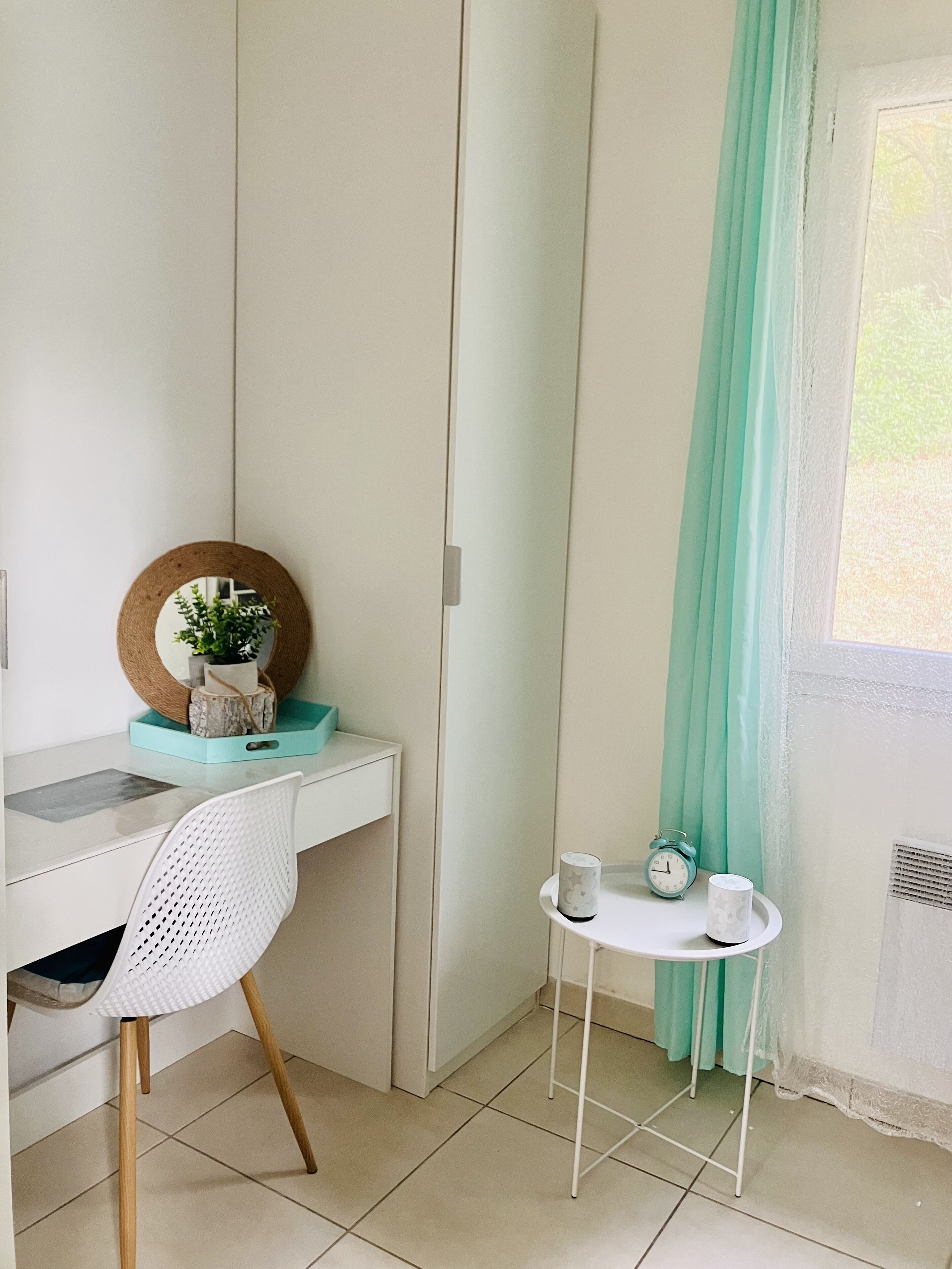 A room with a white desk, a white chair, a small side table with a clock and a cup, and a window with teal curtains.