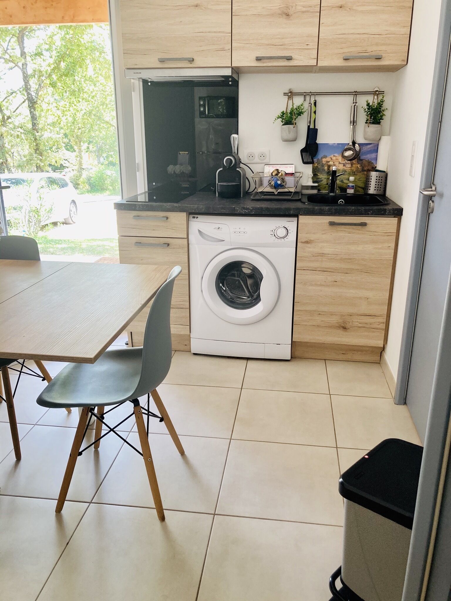 A compact kitchen with a washing machine, a dining table with chairs, and a trash bin.