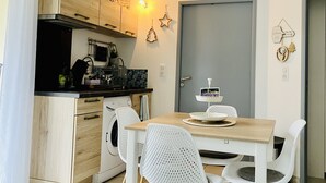 Kompakti keittiö, jossa on puiset kaapit, pesukone sekä ruokailutila, jossa on puinen pöytä ja valkoiset tuolit.