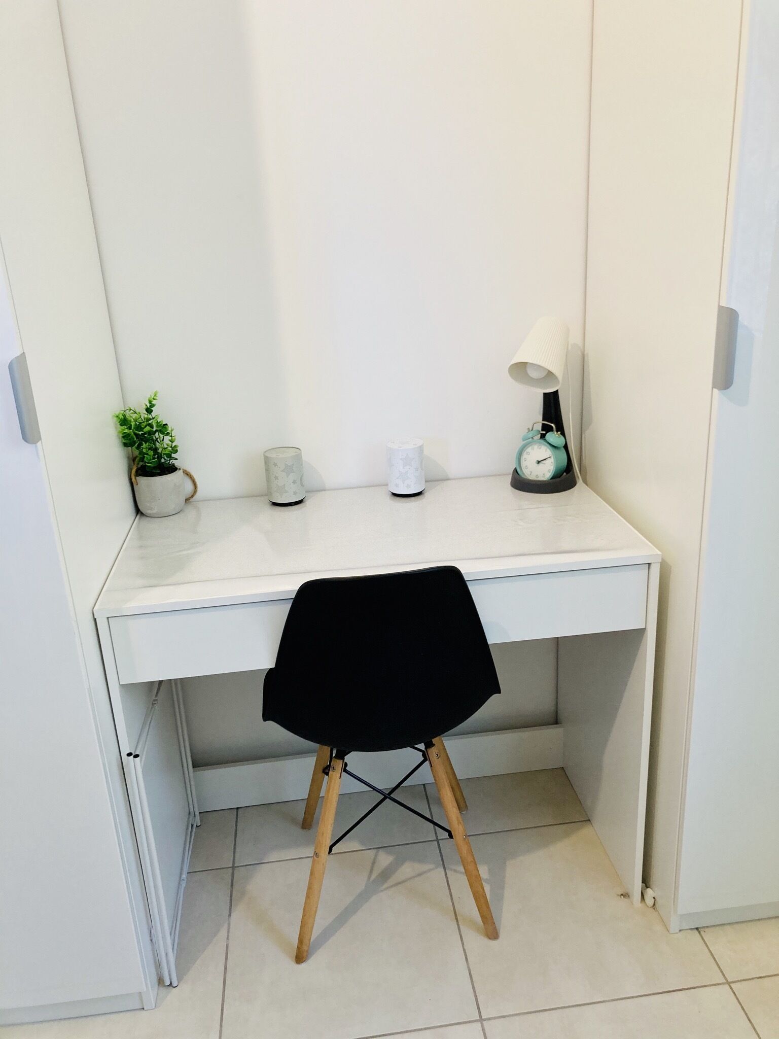 Un bureau blanc avec une chaise noire, une petite plante en pot, une lampe blanche et un réveil turquoise.