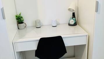Un bureau blanc avec une chaise noire, une petite plante en pot, une lampe blanche et un réveil turquoise.