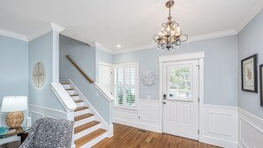 A spacious hallway with a wooden staircase, a chandelier, and a door with a decorative glass panel. - Kappi’s Luxury Coastal Retreat (North Charleston)
