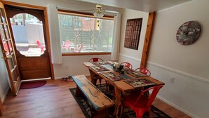 Un comedor con una mesa de madera, sillas rojas y un banco. Se ve una ventana con vistas al exterior, una puerta y una pared con un reloj y un cartel.
