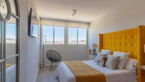 A bedroom with a large bed, a window with a view of the city, a chair, and a lamp.