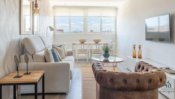 A modern living room with a sofa, coffee table, and TV. There are windows with a view of buildings and a ceiling with a light fixture.