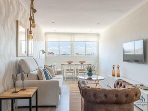 A modern living room with a sofa, coffee table, and TV. There are windows with a view of buildings and a ceiling with a light fixture.