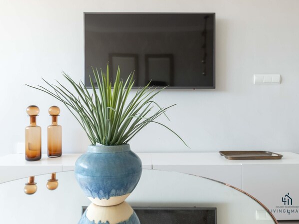 A modern living room with a flat-screen TV mounted on the wall, a blue vase with green plants on a white console table, and two wooden bottles.