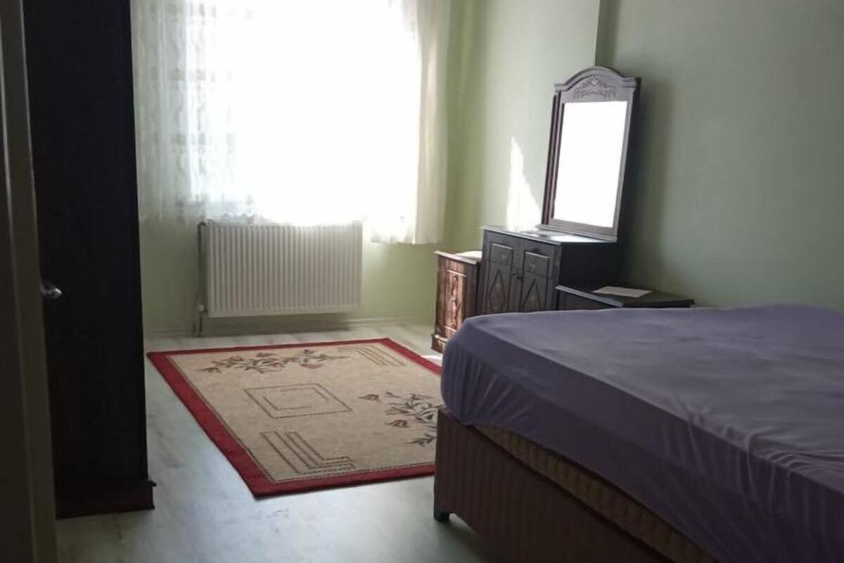 Quarto com cama, cômoda, espelho, radiador e janela com cortinas.