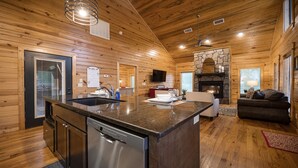 A wooden cabin interior with a kitchen island, a stone fireplace, and a living area with a couch and TV.