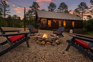A cabin with a fire pit surrounded by Adirondack chairs, a deck, and string lights.