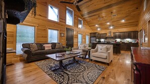 A spacious living area with a dark brown sofa, a beige armchair, and a coffee table. There are large windows and a wooden ceiling.