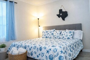 A bedroom with a bed, a wall-mounted Texas flag decoration, a bedside lamp, and a basket with towels. - Clean Pool Walk to Beach - Pet Ok - Ground Floor (South Padre Island)