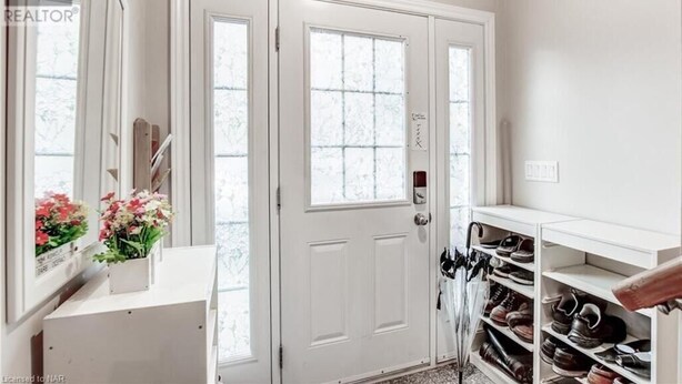 Une porte blanche avec une fenêtre, un porte-chaussures et une petite table blanche ornée de fleurs.
