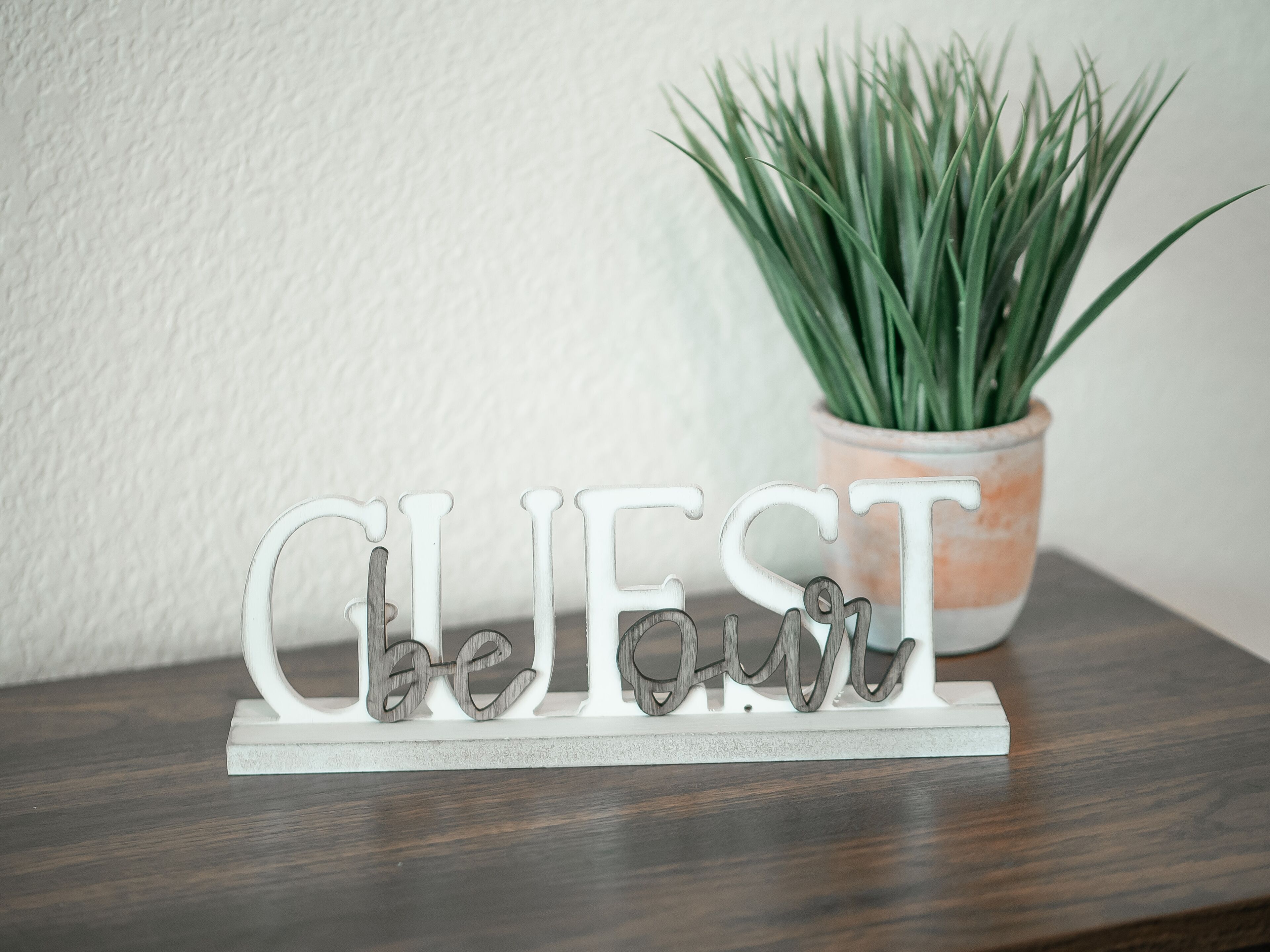 A decorative sign spelling "GUEST" on a white base, placed on a wooden surface with a potted plant in the background.