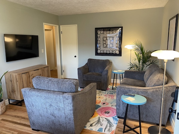 A living room with a flat-screen TV, two grey armchairs, a coffee table, a floor lamp, and a framed artwork on the wall.