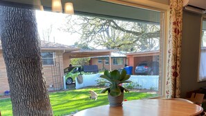 Un comedor con una mesa redonda y cuatro sillas, una planta en maceta y vistas a un patio trasero con valla blanca y coches aparcados.