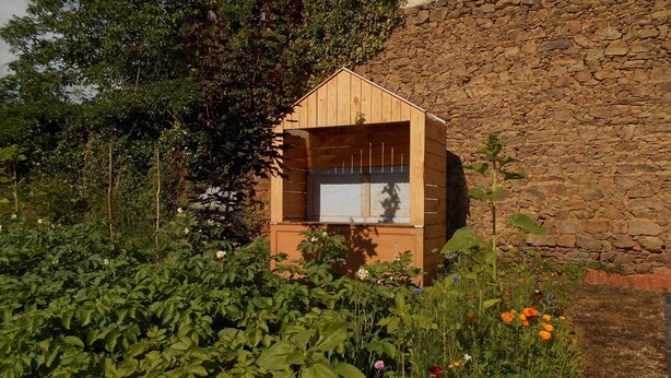 Een houten constructie met een schuin dak, mogelijk een tuinhuisje of een klein huis, omgeven door weelderige begroeiing en een stenen muur.