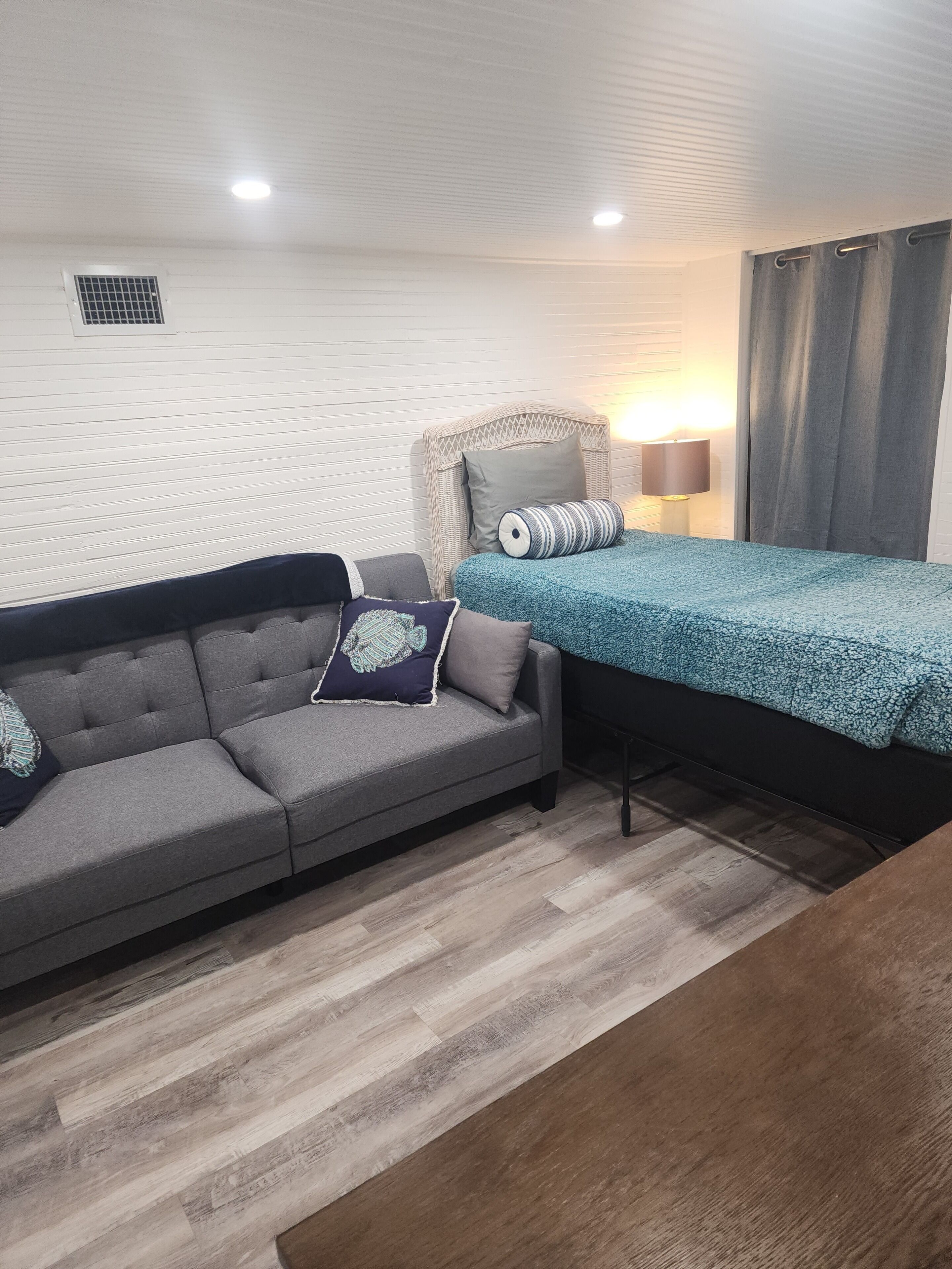 Une pièce avec un canapé gris, un lit recouvert d’une couvre-lit à motifs bleus, une chaise en osier et une lampe.