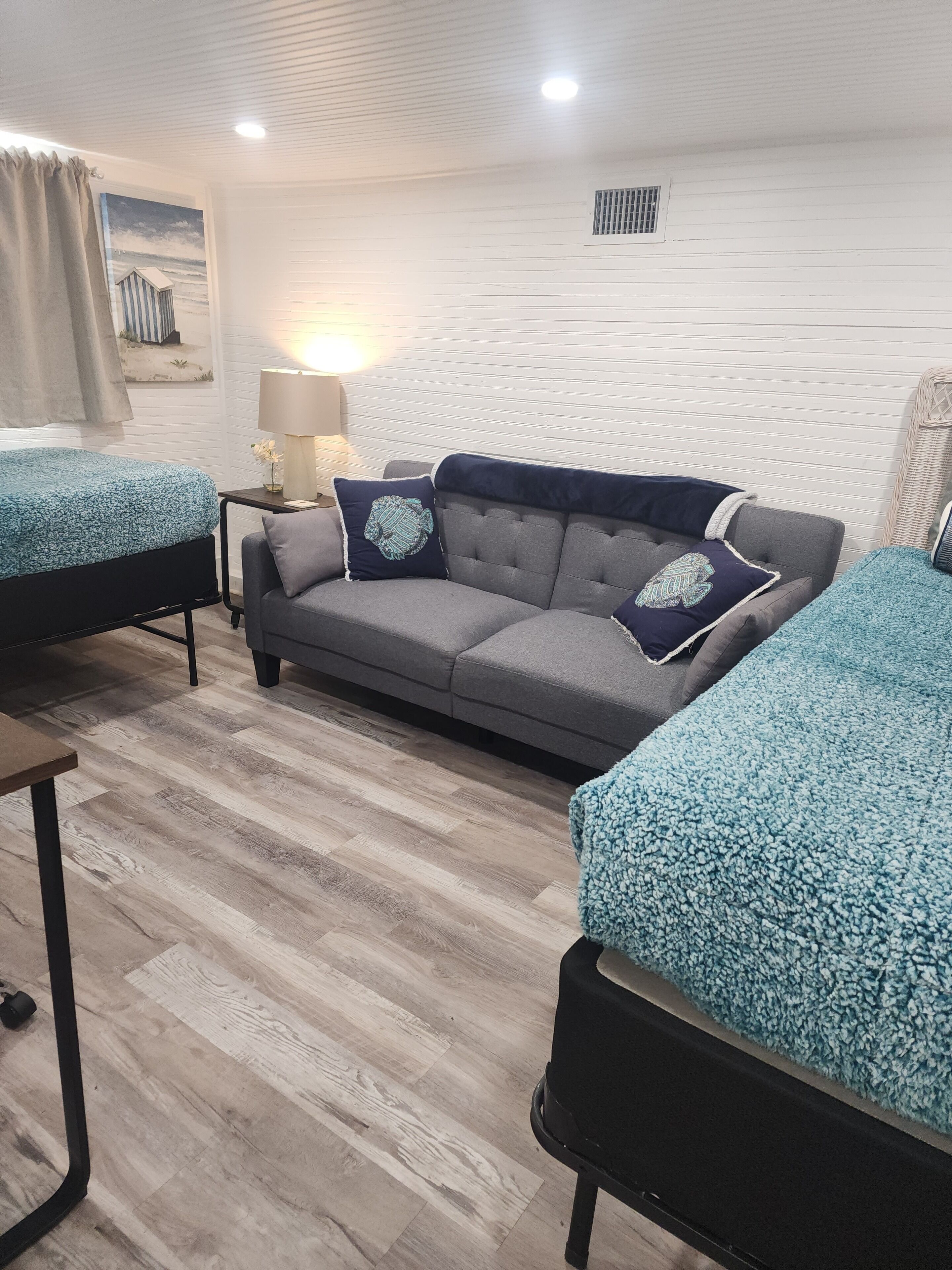 Um quarto com um sofá cinzento, duas camas de solteiro com roupa de cama azul, uma mesinha de centro com um abajur e um respiradouro fixado na parede.