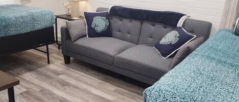 Um quarto com um sofá cinzento, duas camas de solteiro com roupa de cama azul, uma mesinha de centro com um abajur e um respiradouro fixado na parede.