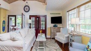 A living room with a sofa, armchair, and a television. There is a door with a sign that reads "IN LOVE BY THE SEA".