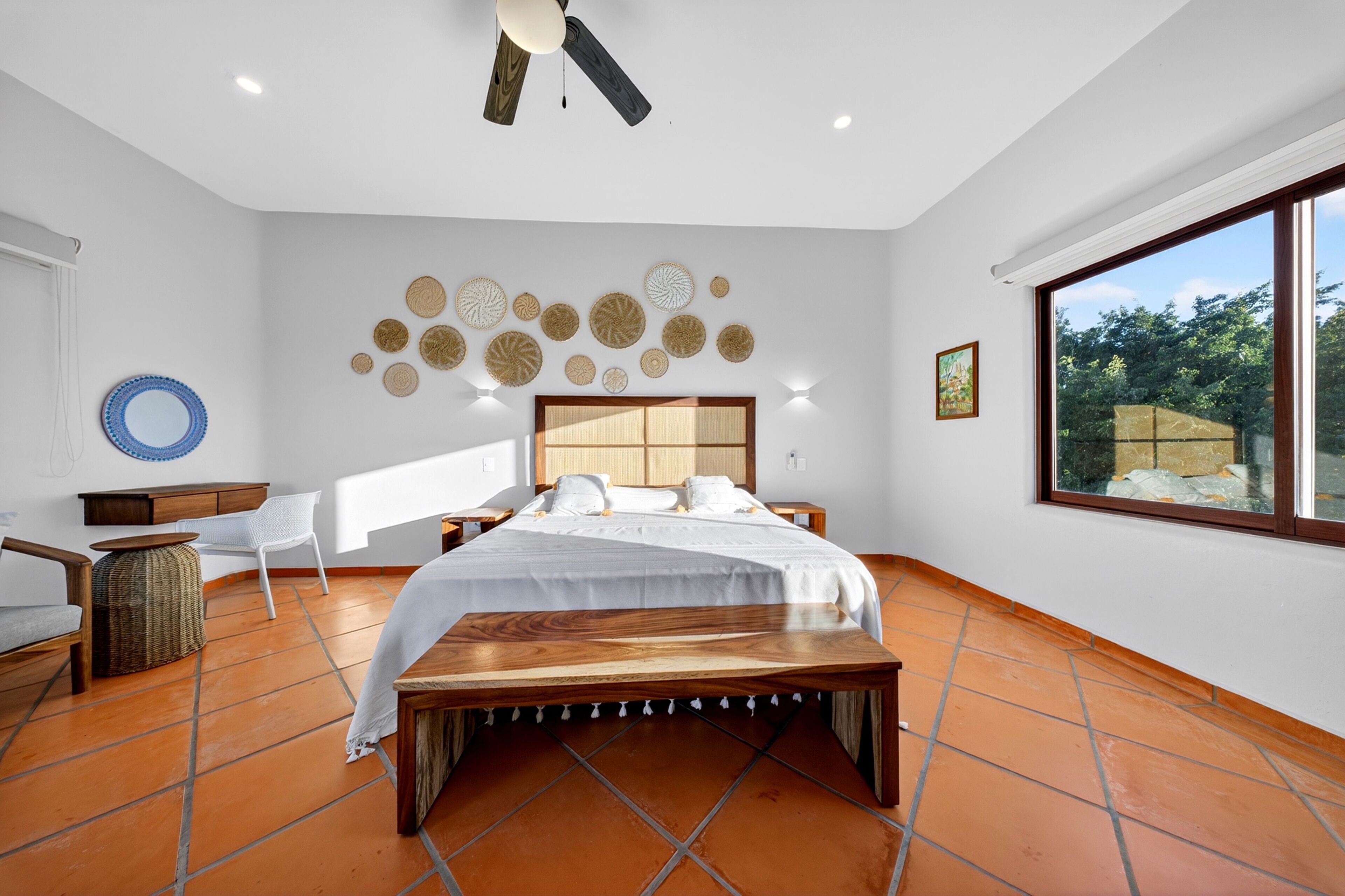 A bedroom with a large bed, a ceiling fan, a view of trees, and a tiled floor.