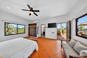 A bedroom with a bed, a ceiling fan, a TV, and large windows.