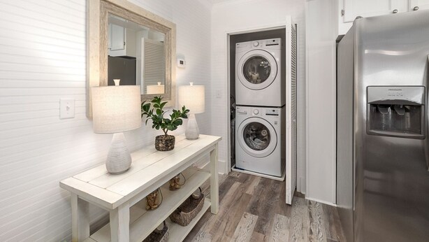 A modern laundry room with two stacked front-loading washers, a stainless steel refrigerator, and a white console table with two lamps and a potted plant.