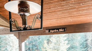 Une terrasse en bois avec une table ronde et quatre chaises, une lampe suspendue et un panneau indiquant « Après-ski ».