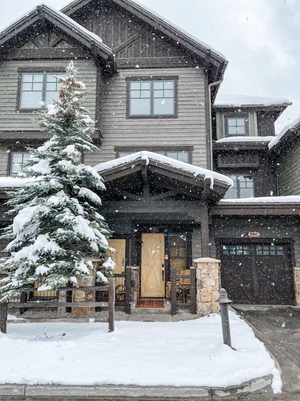 Una casa de madera de dos pisos con un techo cubierto de nieve y un abeto grande en el frente.