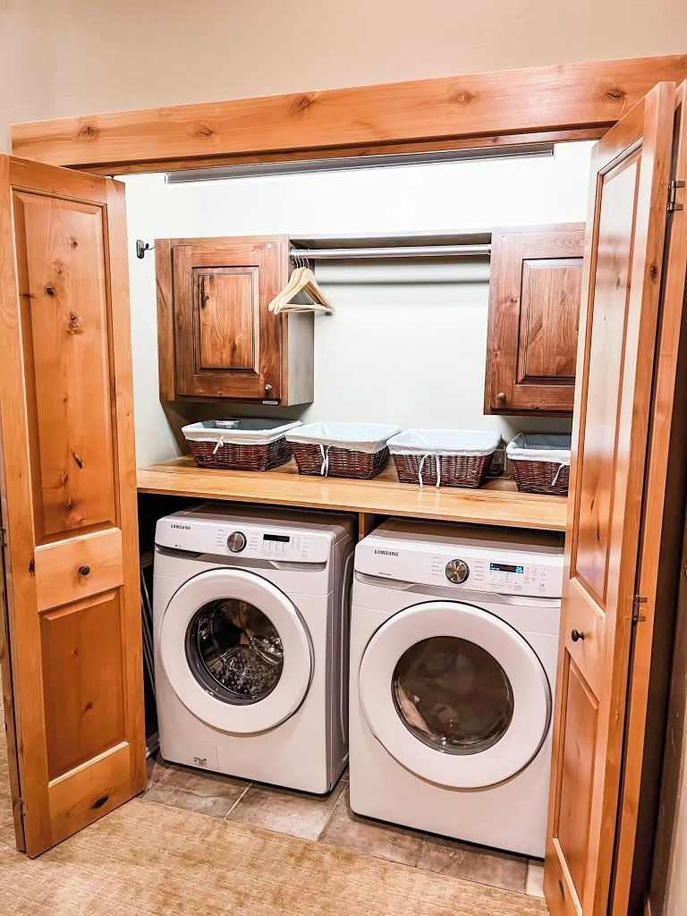 A compact laundry area with two washing machines, wooden cabinets, and hanging baskets.