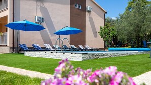 Ein Poolbereich mit Liegestühlen und blauen Sonnenschirmen, eine Steinmauer und ein Gebäude mit Klimaanlagen.