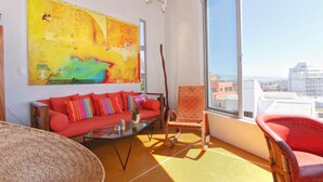 Ein modernes Wohnzimmer mit einem großen abstrakten Gemälde, einem roten Sofa, einem Holzstuhl und Blick auf die Stadt durch große Fenster.