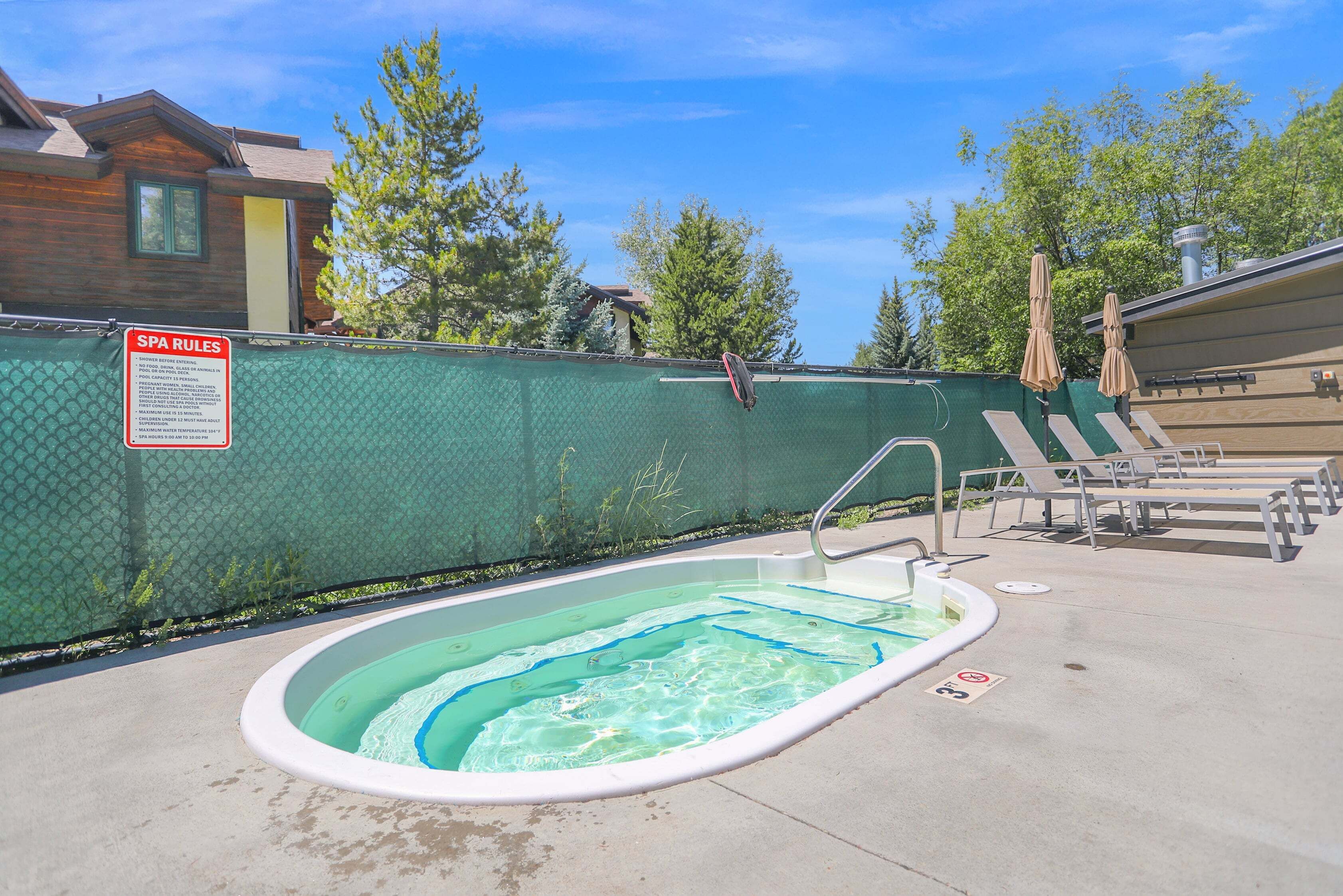 An outdoor swimming pool with a spa area, lounge chairs, and a fence with a sign.