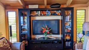 A living room with a large flat-screen TV on a black entertainment unit, surrounded by bookshelves and decorative items. - Luxury Condo in Wailea with sweeping views (Kihei)
