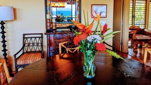 A dining area with a wooden table, chairs, a vase with flowers, a chandelier, and a view of the ocean through a window. - Luxury Condo in Wailea with sweeping views (Kihei)